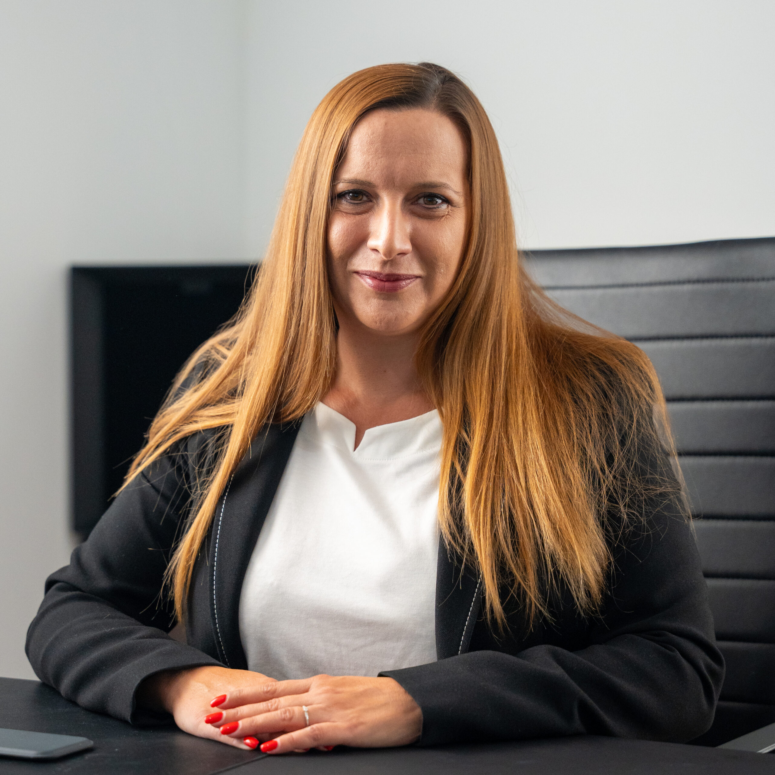 eine frau mit langem glattem kastanienbraunem haar, die ein weißes oberteil und einen schwarzen blazer trägt, sitzt mit gefalteten händen an einem schreibtisch. sie lächelt leicht in einer büroumgebung.