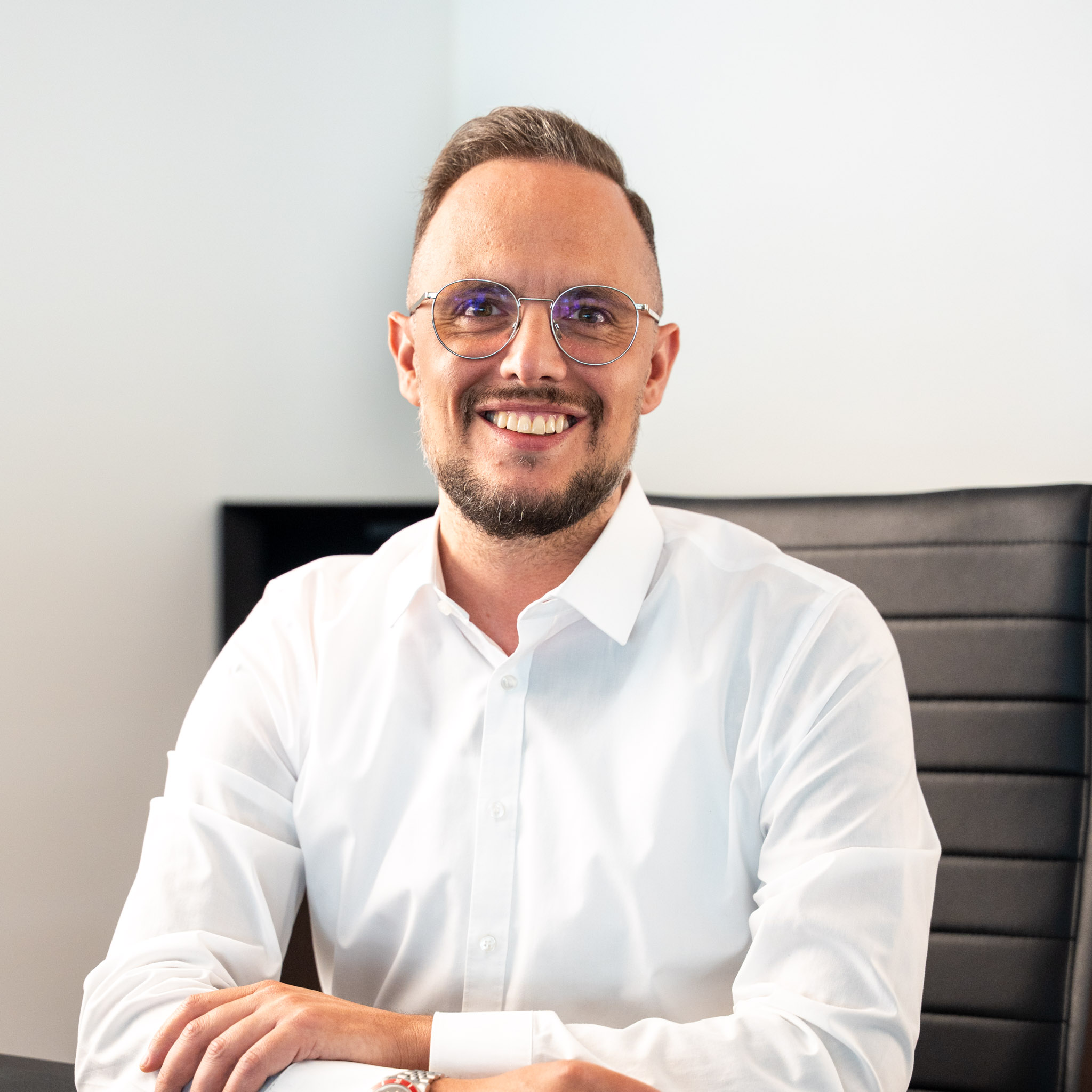 ein mann mit kurzen haaren, einem gestutzten bart und einer brille sitzt lächelnd an einem schreibtisch. er trägt ein weißes hemd und sitzt in einem modernen schwarzen bürostuhl vor einem hellen hintergrund.