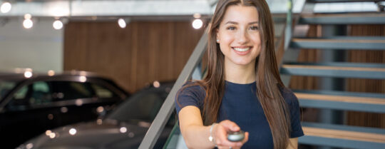 Eine lächelnde Frau mit langen braunen Haaren hält einen Autoschlüsselanhänger in der Hand. Sie steht in einem Innenraum in der Nähe einer Treppe, und im Hintergrund sind Autos zu sehen, was auf einen Autosalon hindeutet.